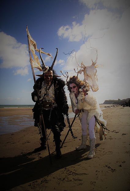 Two individuals on the beach dressed as traditionally costumed characters from folklore, one ont he left holding a tattered yellow flag and wearing a skull-like mask with horns, and holding a chain. The individual on the right is dressed in white with antlers and is holding a horse skull on a rod. There are bright skies and clouds in the background.