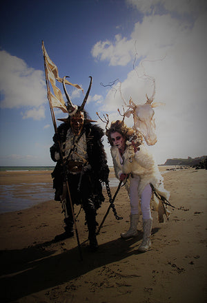 Two individuals on the beach dressed as traditionally costumed characters from folklore, one ont he left holding a tattered yellow flag and wearing a skull-like mask with horns, and holding a chain. The individual on the right is dressed in white with antlers and is holding a horse skull on a rod. There are bright skies and clouds in the background.